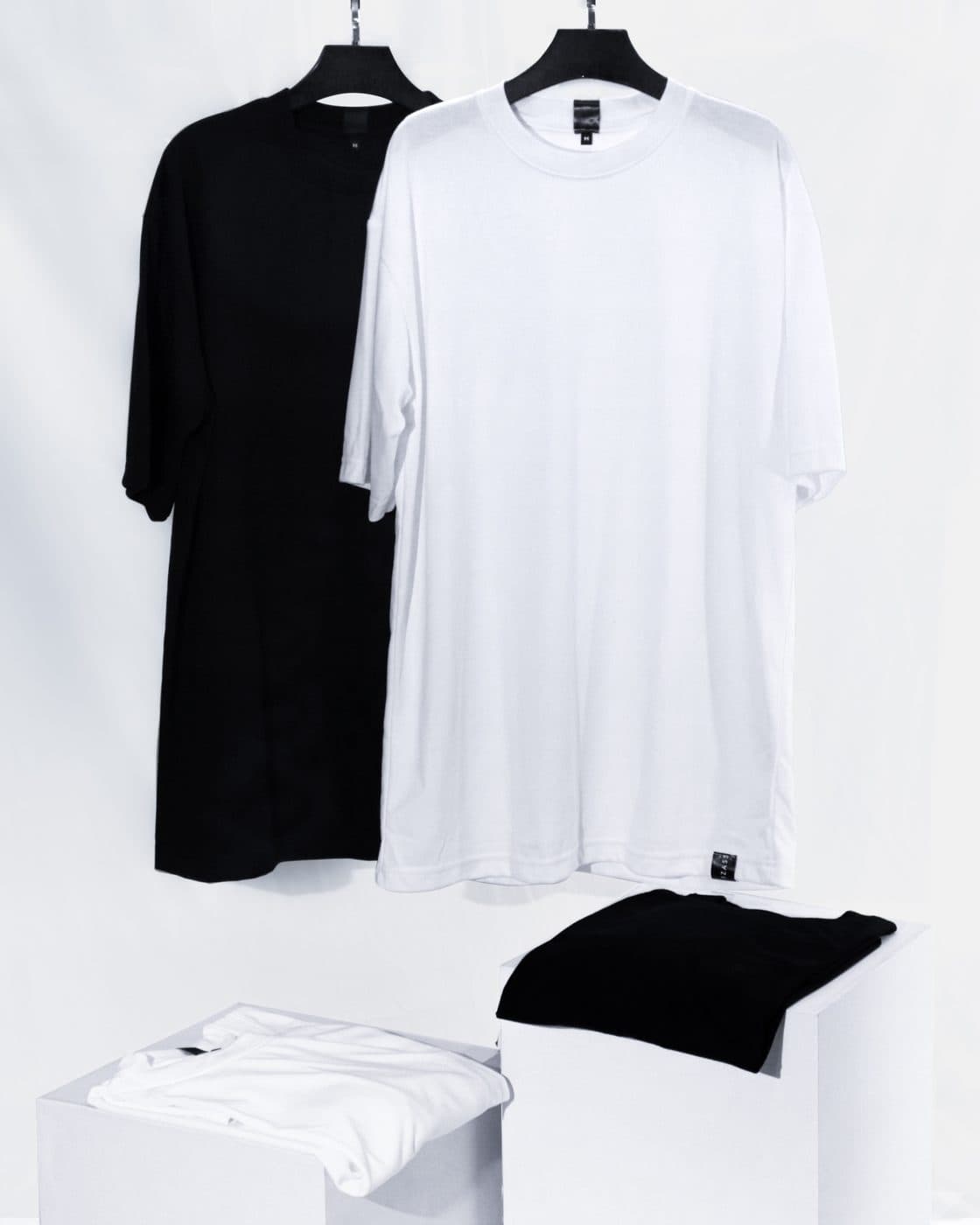 เสื้อยืดโอเวอร์ไซซ์สีดำและสีขาว แขวนบนไม้แขวนและพับวางบนกล่อง
